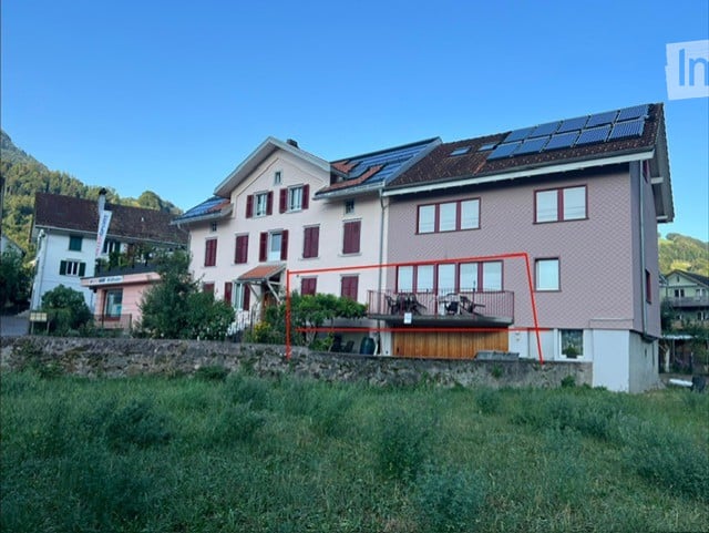 Two-story residential house with solar panels on the roof, a balcony with outdoor furniture, and a stone retaining wall in front.