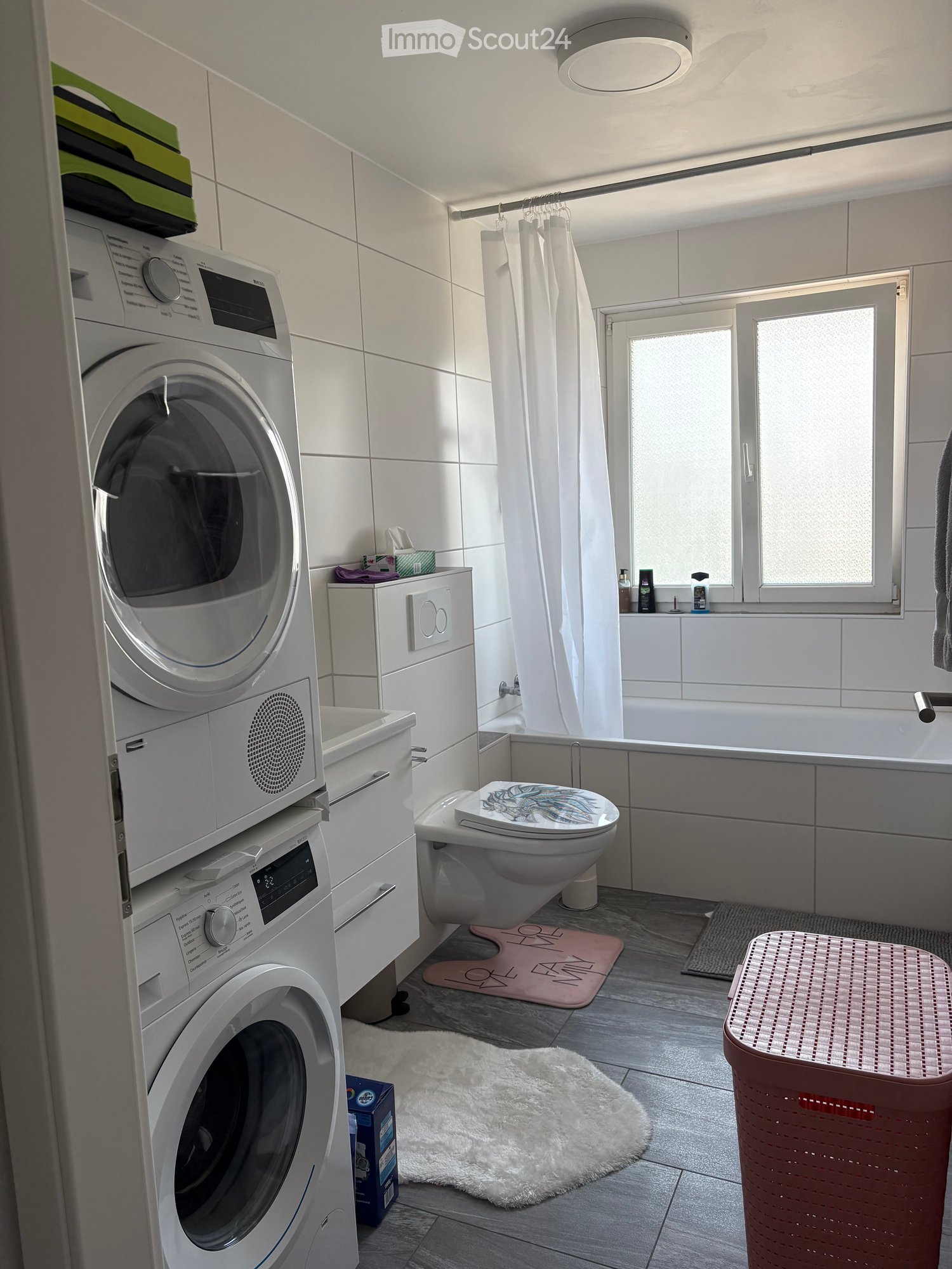 Modern bathroom with white tiled walls, a bathtub, toilet, washing machine, and a shower area with a curtain. Includes a small cabinet, a fluffy rug, and a laundry basket.