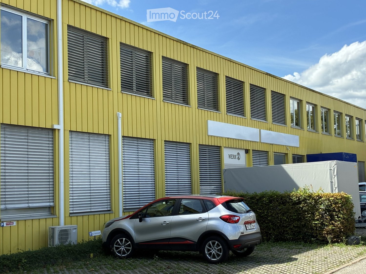 Yellow building with multiple windows and blinds, a silver car parked in front, and a sign reading "WERK 2" on the wall.