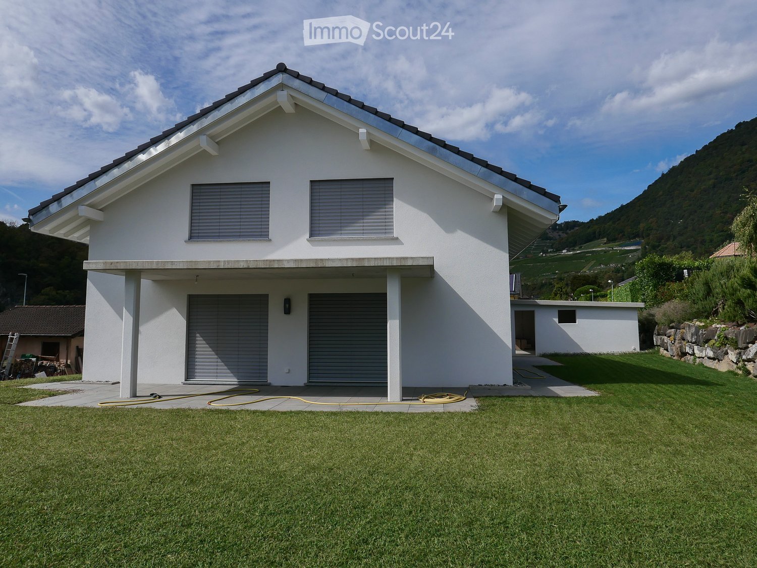 Modern two-story house with white exterior, garage, and large windows, situated on a grassy lawn with a scenic mountain backdrop.