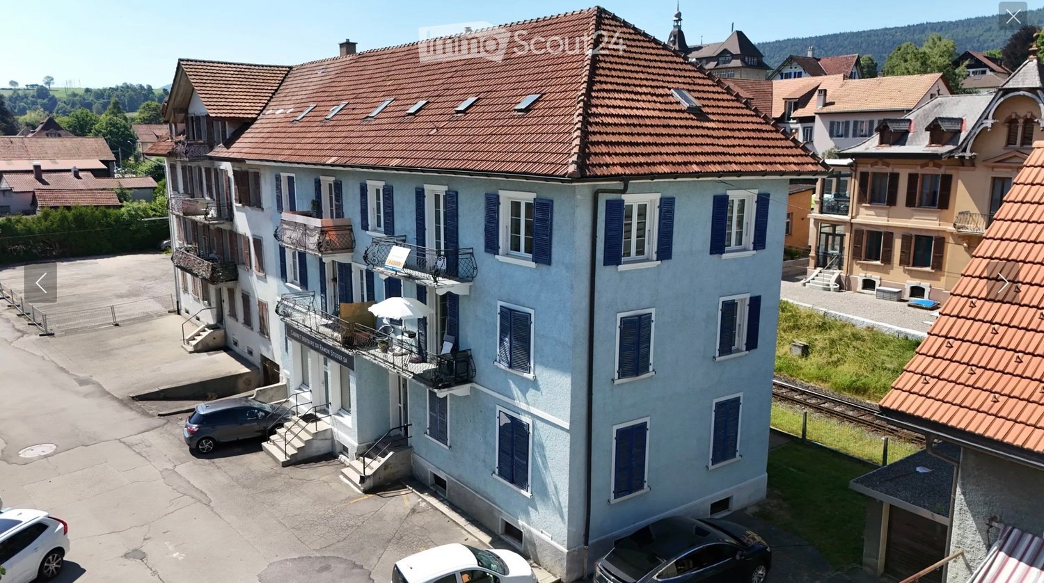 Multi-story residential building with blue shutters, balconies, and a red-tiled roof, surrounded by other houses and parked cars.