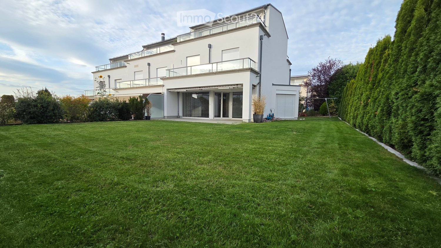 Well-maintained lawn in front of a modern two-story house with white exterior, large windows, and a balcony on the upper level.