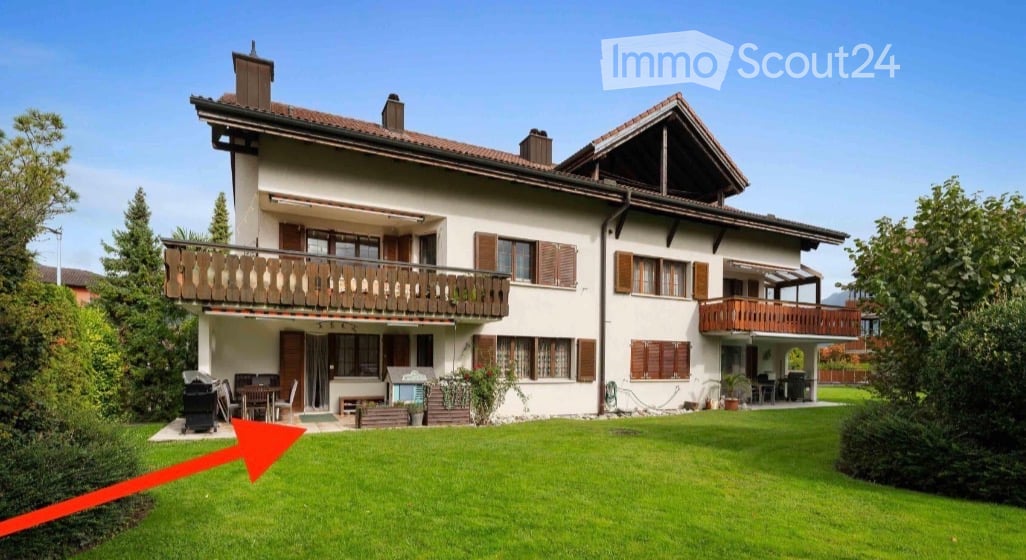 Two-story house with white walls, brown shutters, and balconies on both floors. The house features a well-maintained lawn and outdoor seating area.