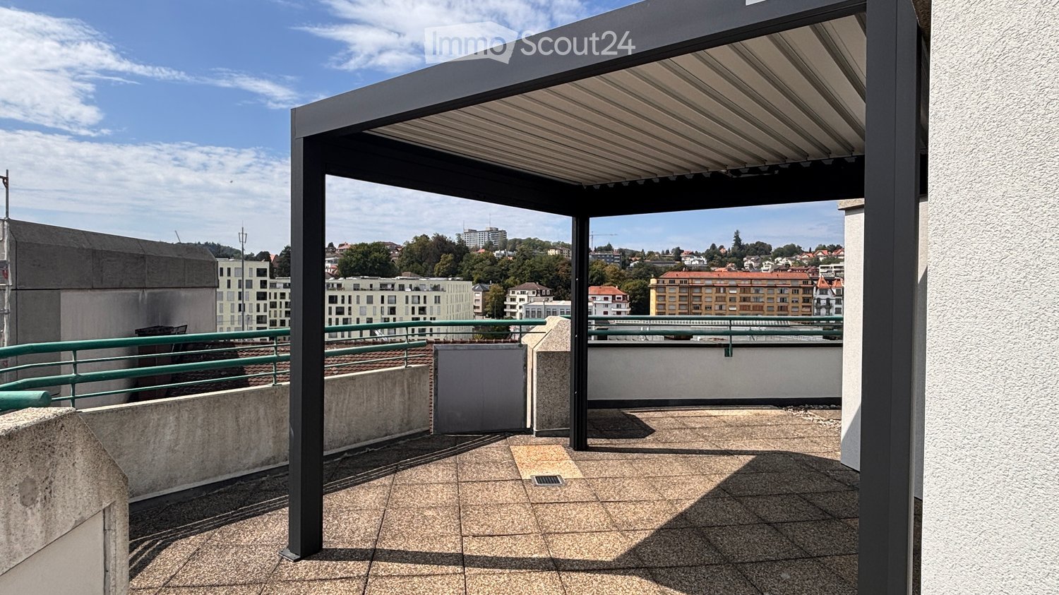Modern rooftop terrace with a retractable awning, tiled floor, green metal railing, and a view of the cityscape.