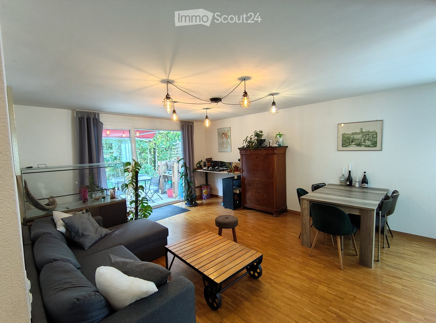 Bright living room with wooden flooring, a grey sofa, a wooden coffee table, a dining table with chairs, a wooden cabinet, and a desk area with a computer.