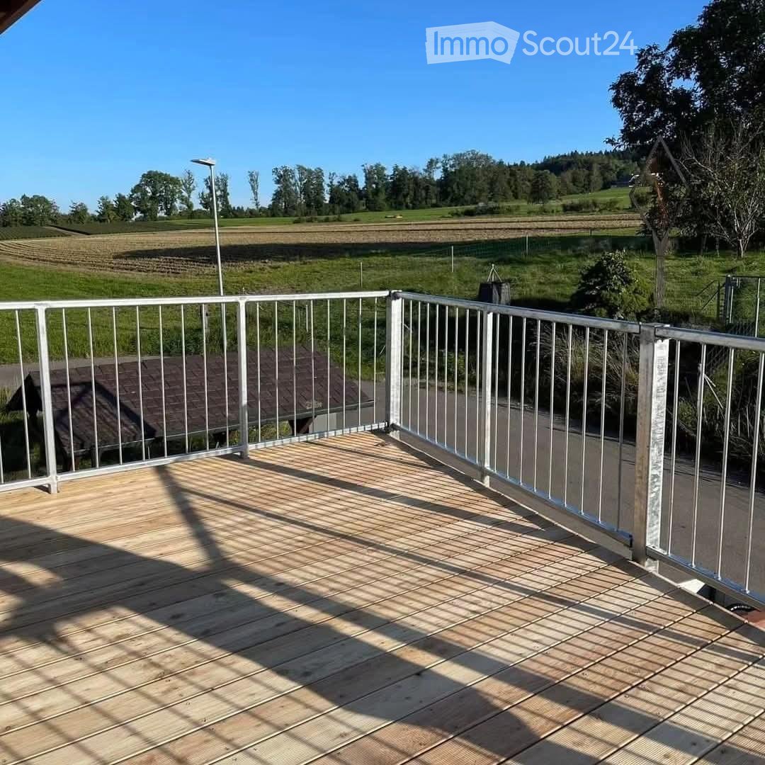 Wooden deck with metal railing, overlooking a green field and trees under a clear blue sky.