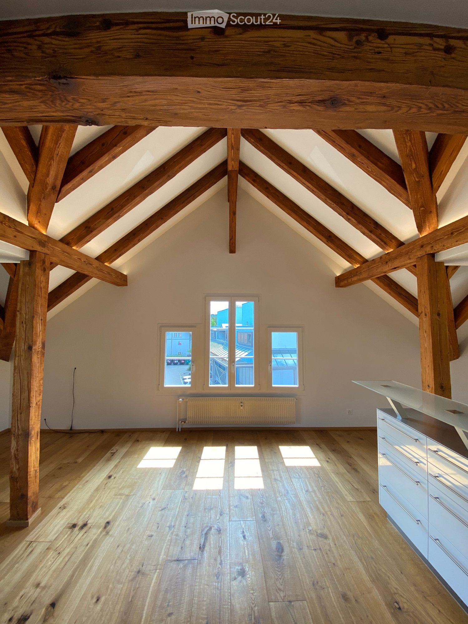 Bright, airy attic space with exposed wooden beams, wooden flooring, and large windows allowing natural light. A modern white cabinet is visible on the right side.