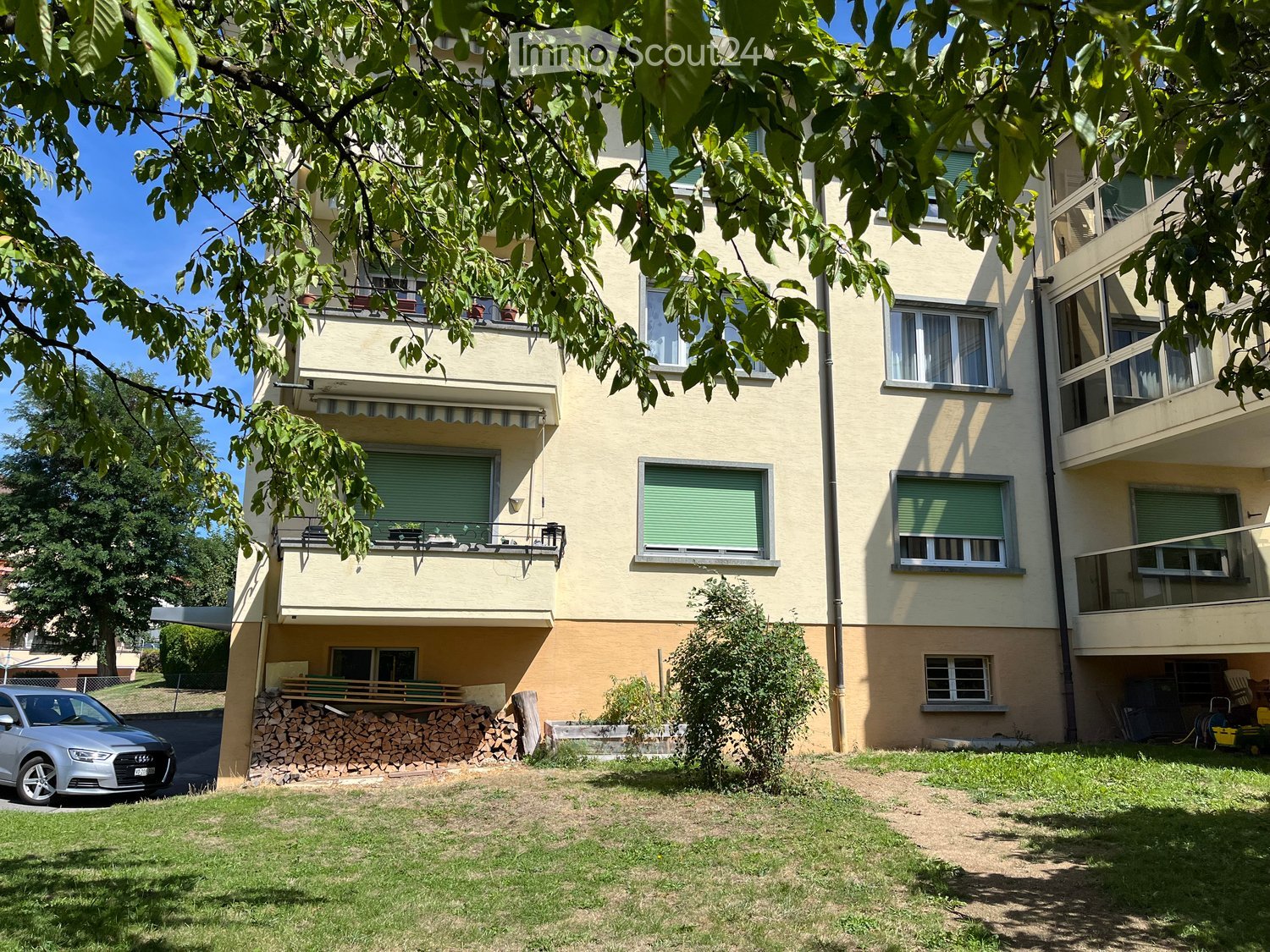 Four-story residential building with balconies, green shutters, and a small garden area with a stack of firewood and a parked car.