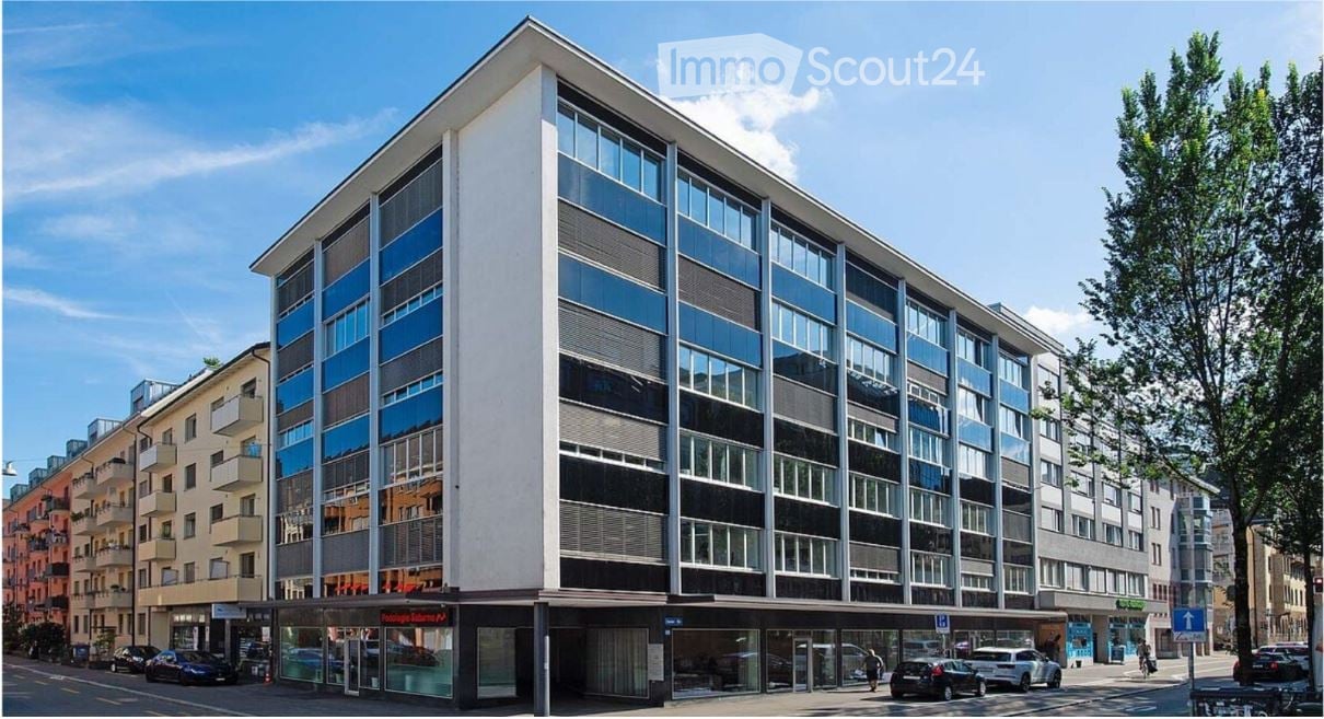 Modern multi-story building with large glass windows and balconies, located in an urban setting with surrounding structures and parked cars.