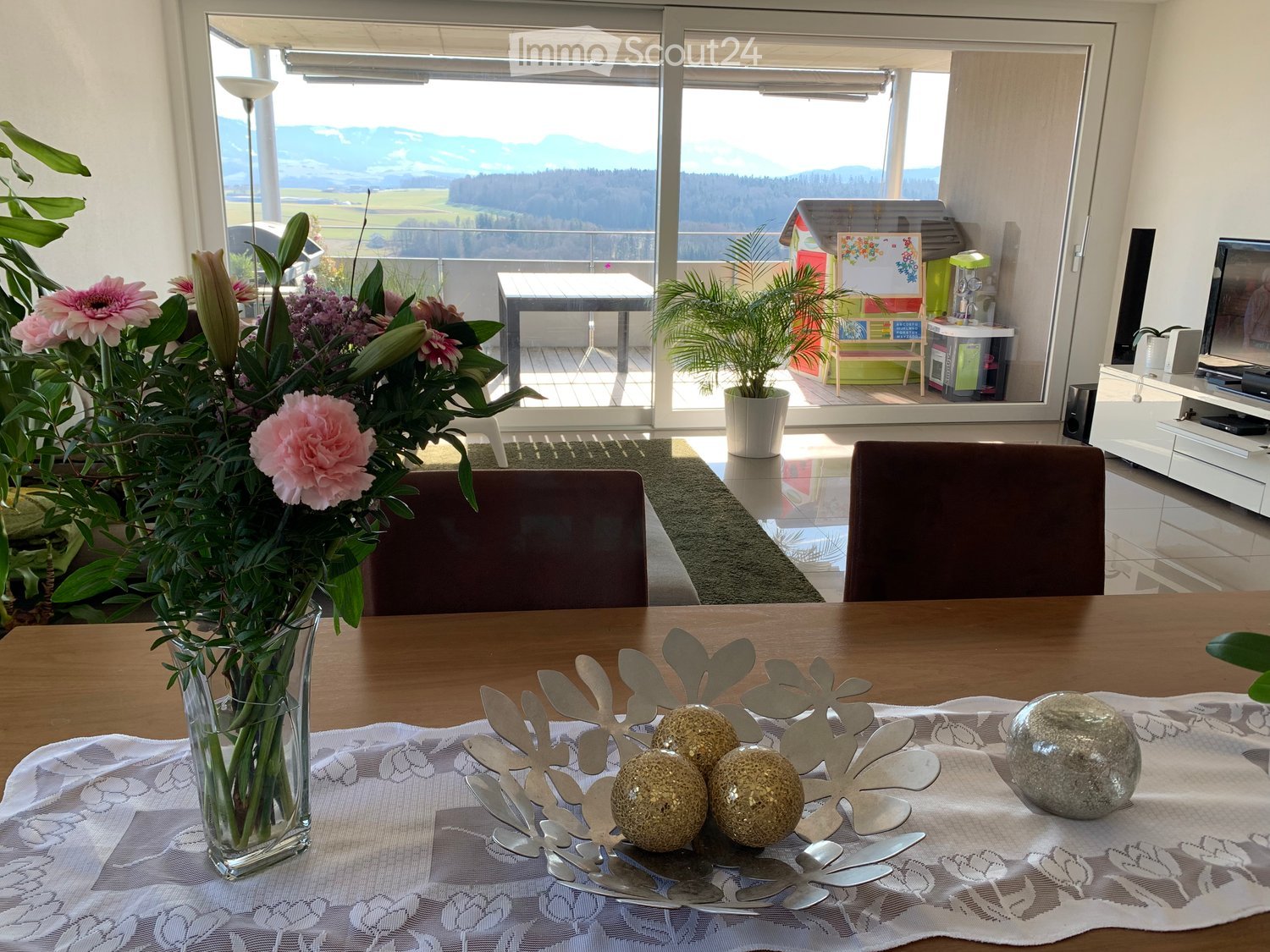 Bright living room with large windows offering a scenic mountain view, featuring a wooden dining table with a floral centerpiece, chairs, a television, and potted plants.
