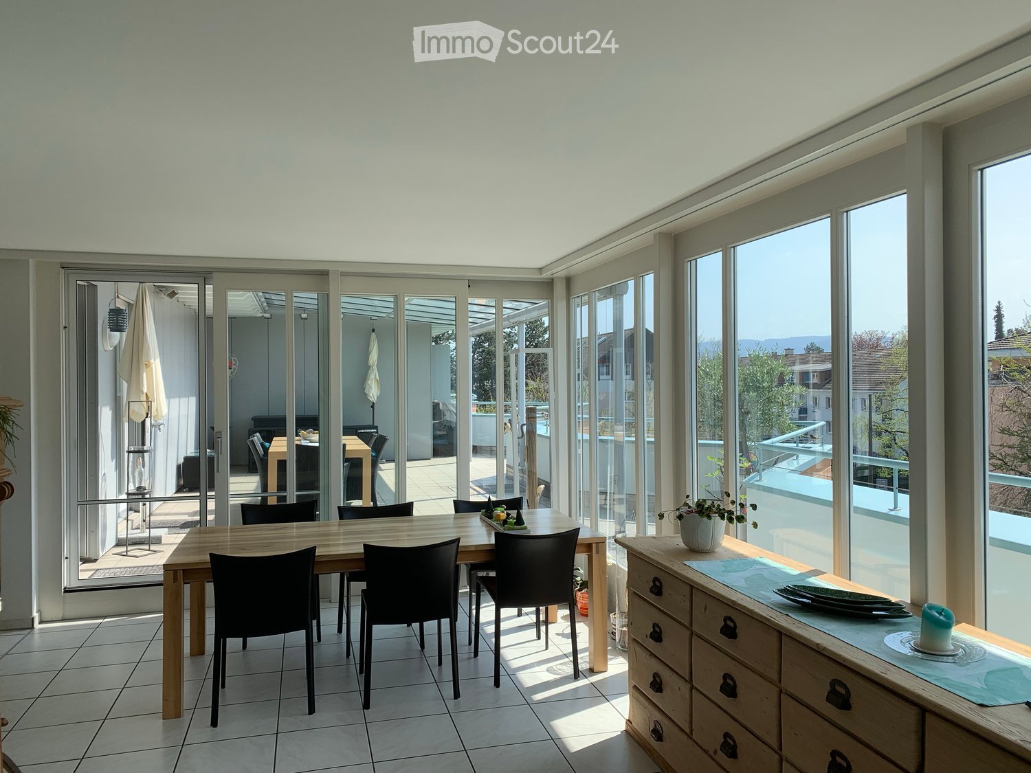 Bright, modern dining area with large windows and sliding glass doors leading to a patio. Features a wooden dining table with black chairs, a sideboard with a candle and vase, and an umbrella stand near the entrance.