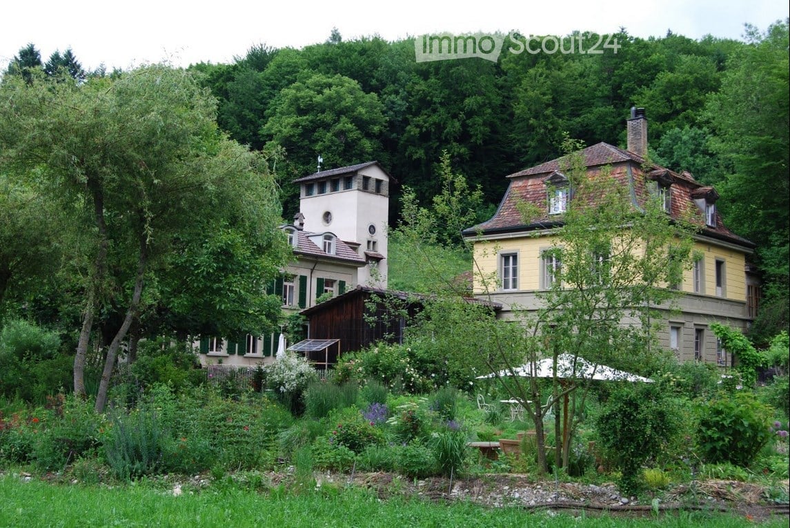 A lush garden area with two historic buildings in the background, surrounded by trees and greenery.
