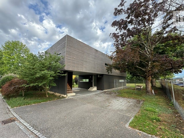 Modern two-story building with a raised ground floor, grey facade, and a small staircase leading to the entrance. The building is surrounded by trees and a paved driveway.