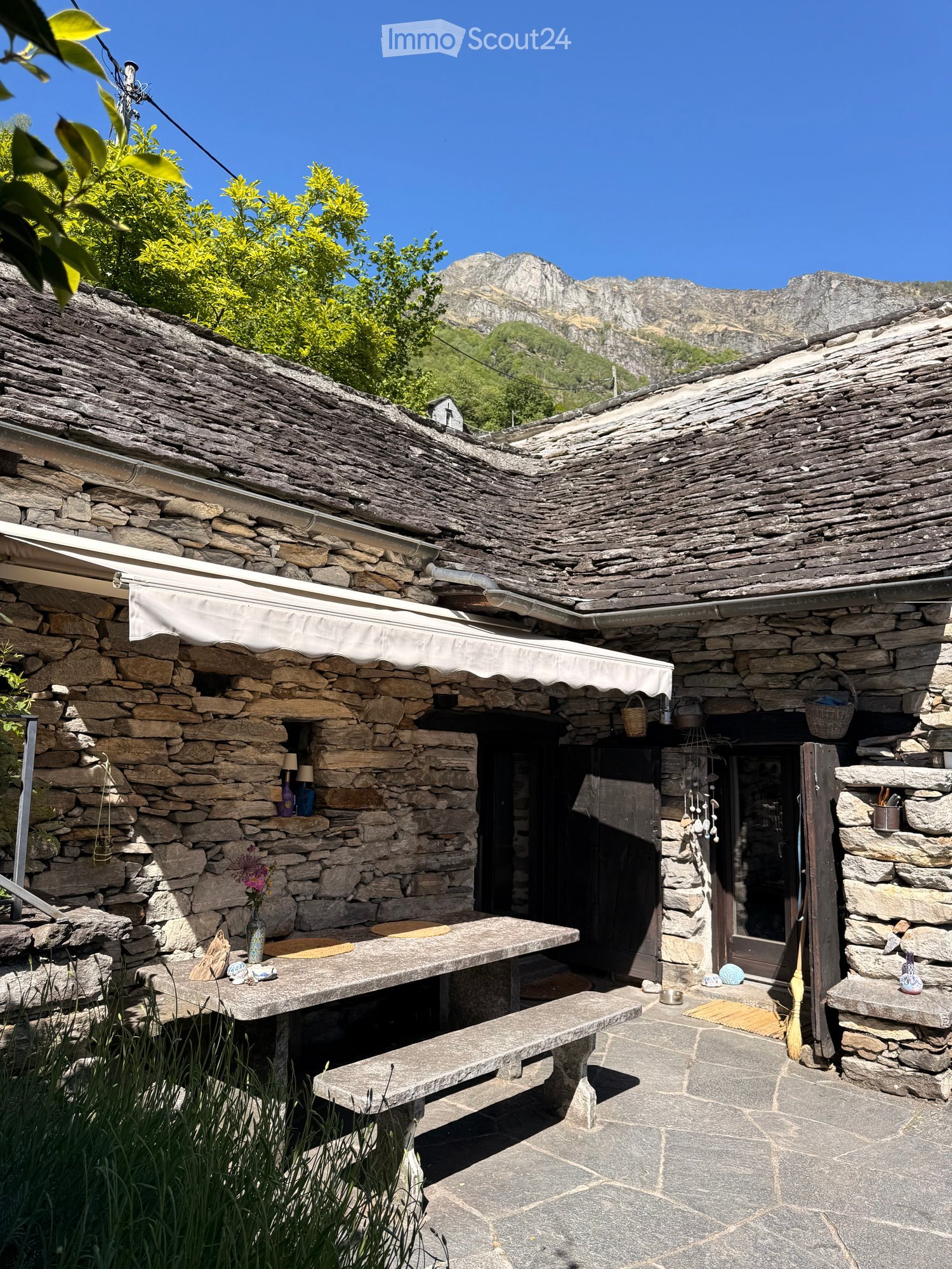 Stone outdoor seating area with a table, bench, vase with flowers, and baskets, set against a stone building with a view of mountains.