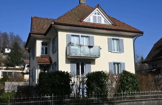 Two-story house with a cream-colored facade, gray shutters, and a balcony on the second floor. The roof is tiled with brown shingles, and the house is surrounded by greenery and a low fence.