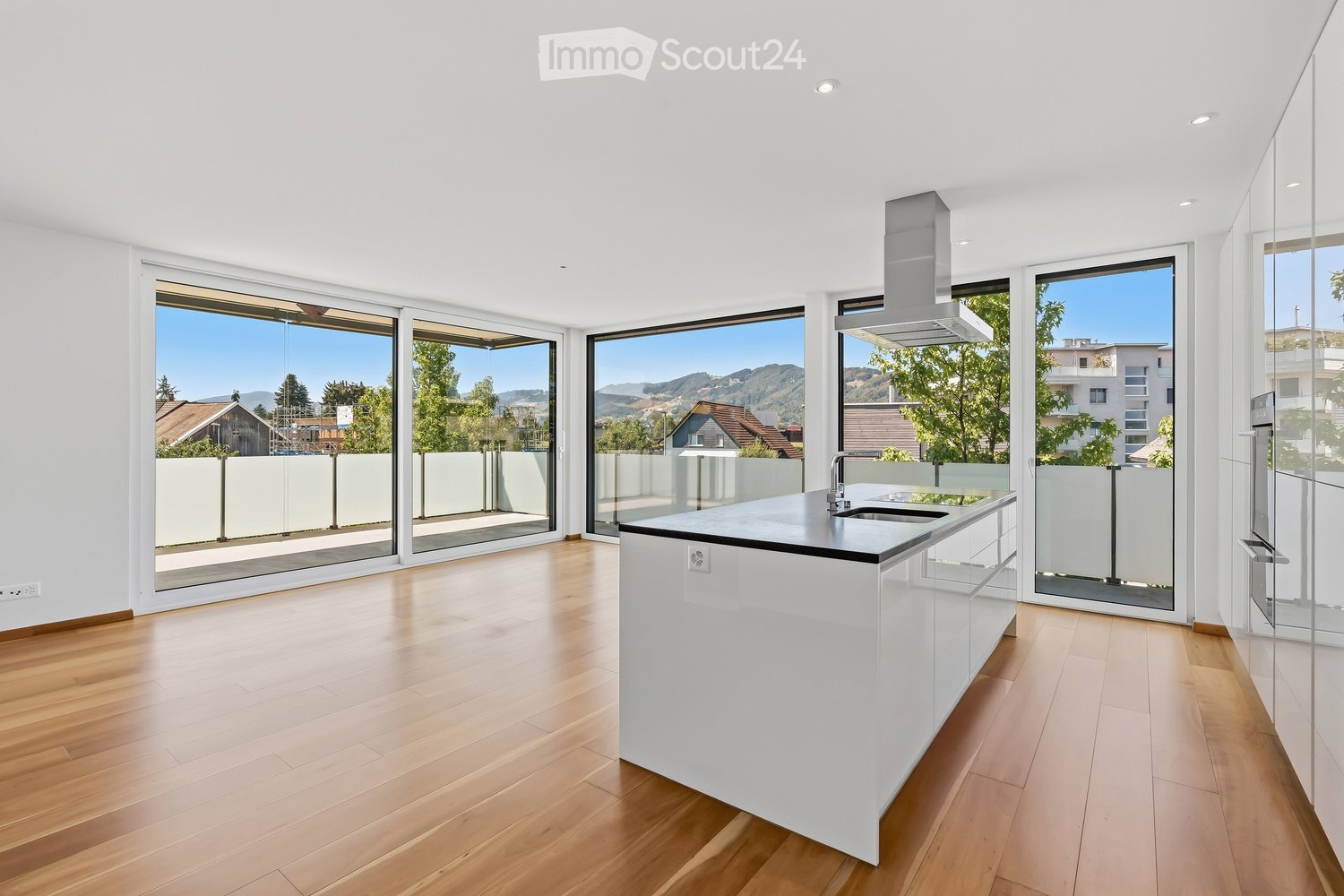 Modern kitchen with white island, wooden flooring, large windows offering a scenic view, and a range hood.