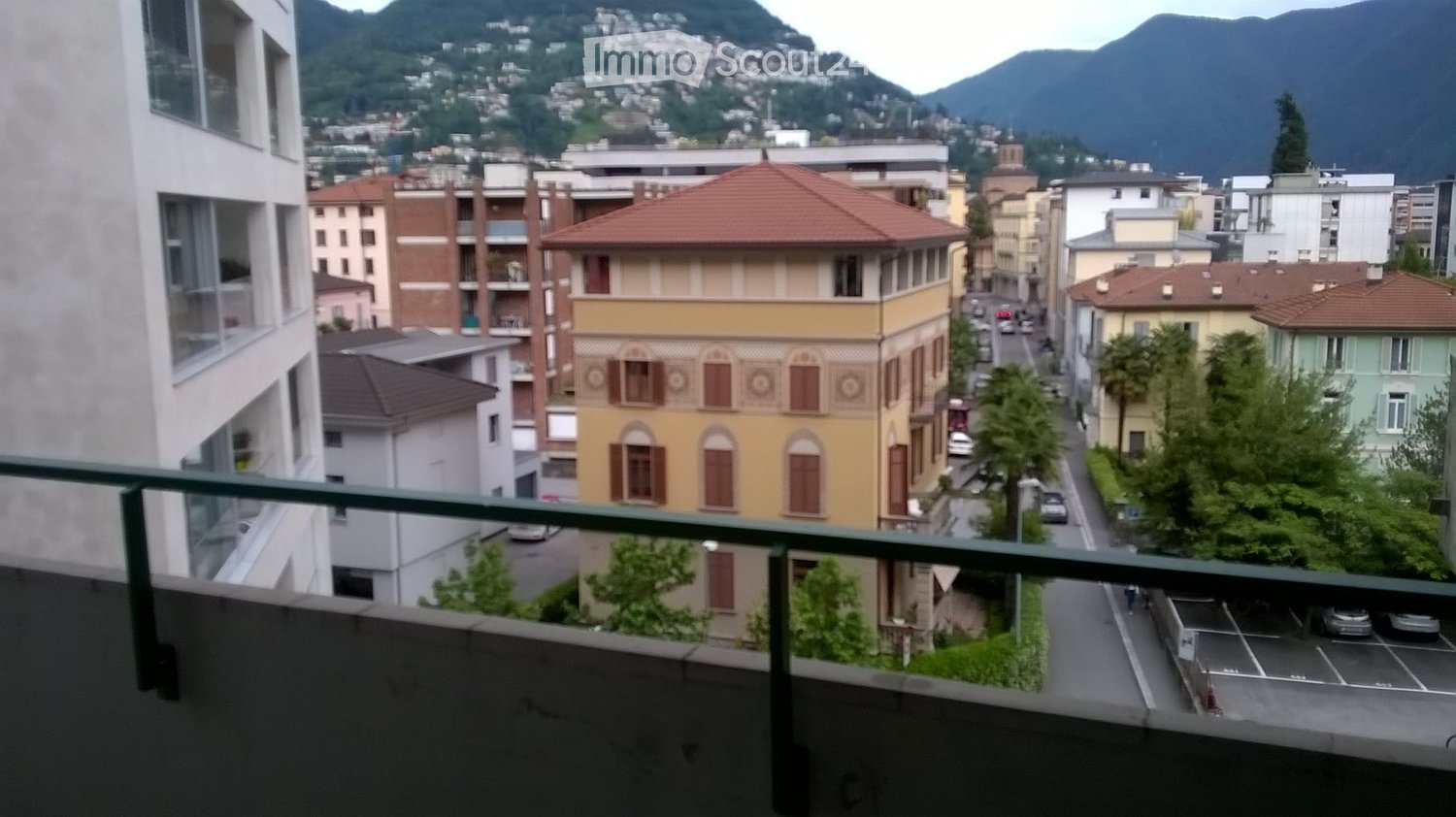 Balcony overlooking a city street with colorful buildings, parked cars, and distant mountains.