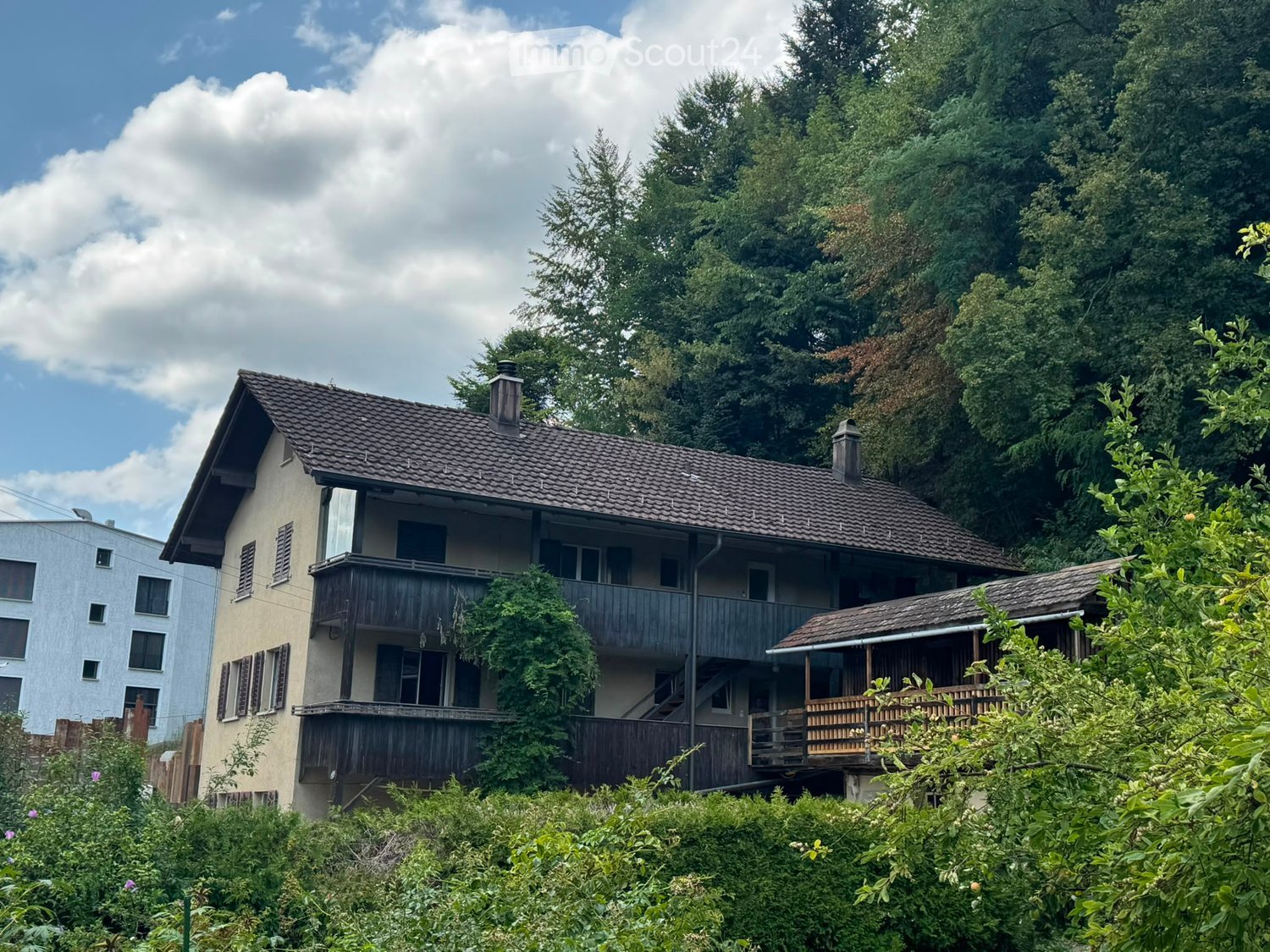 Two-story house with a tiled roof, multiple windows, and wooden balconies surrounded by lush greenery and trees.