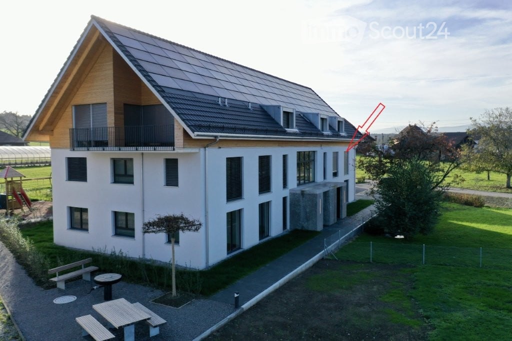 Modern two-story building with white walls, large windows, and a balcony on the upper floor. The roof features solar panels and a wooden facade section. The surrounding area includes a grassy lawn, a small tree, and outdoor seating with benches and tables