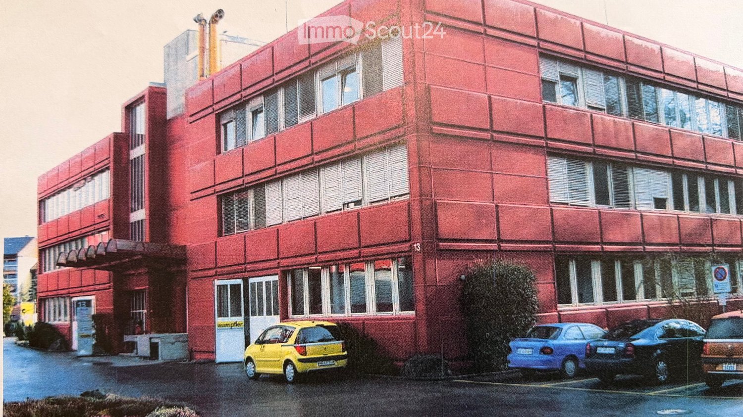 A red multi-story building with numerous windows and balconies, surrounded by parked cars.