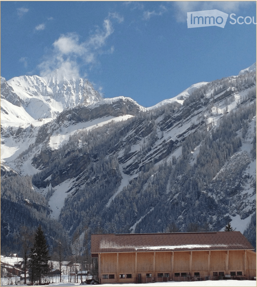 A wooden building with a brown roof and snow-covered ground, surrounded by snow-covered mountains and trees.