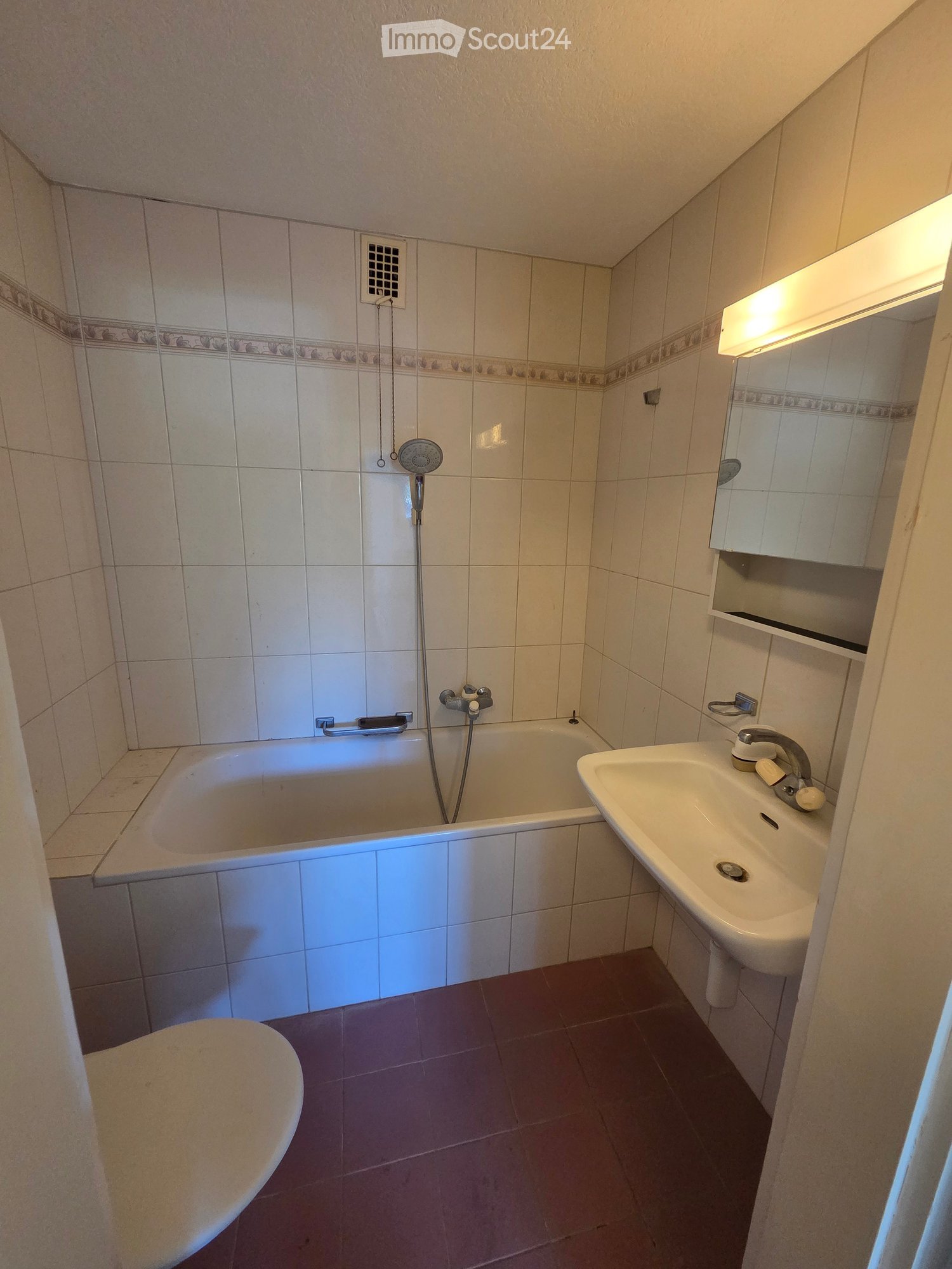 Bathroom with a bathtub, sink, toilet, mirror, and showerhead. The walls are tiled with a decorative border, and the floor is tiled in a dark color.