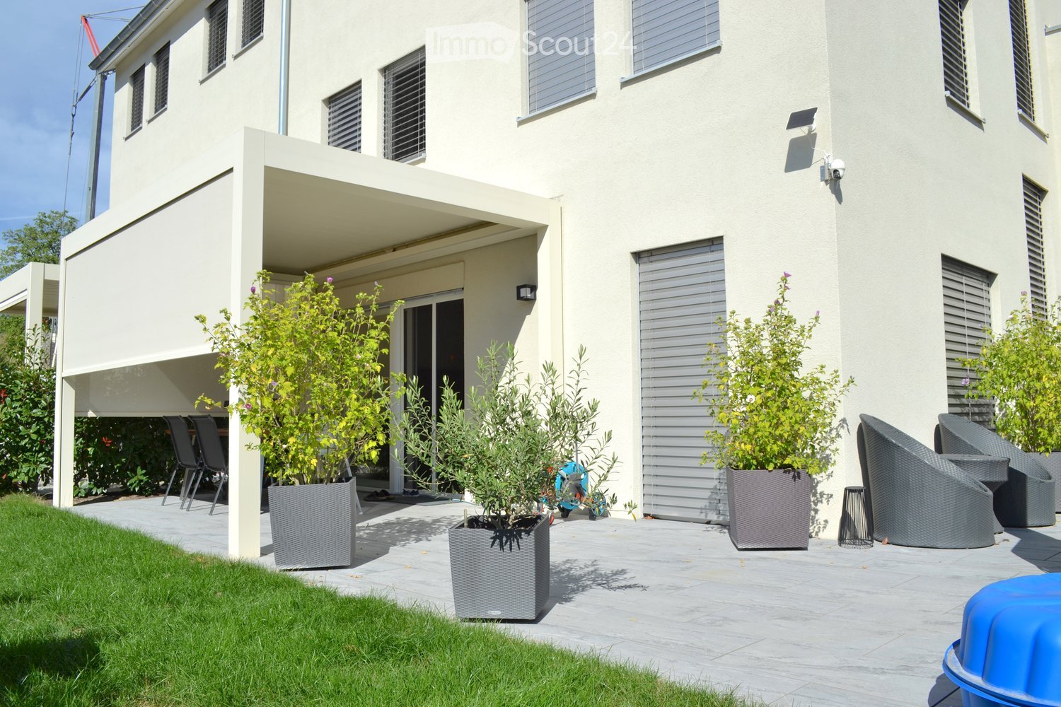 Modern outdoor patio area with grey wicker chairs, a table, and several potted plants, adjacent to a two-story white house with large windows and shutters.