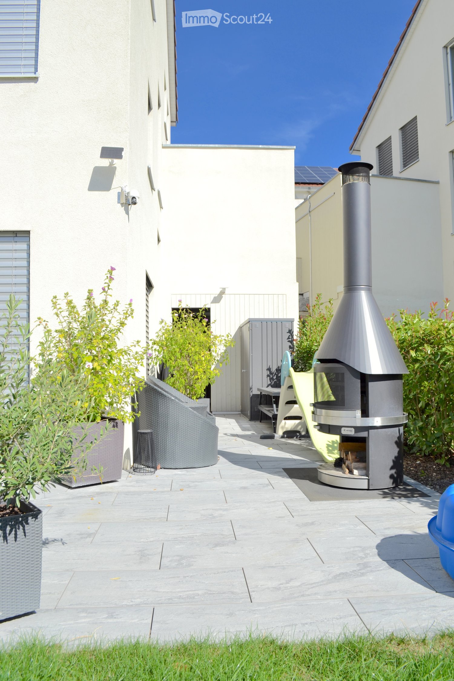 Modern patio with a stone floor, a round wicker chair, a small table, and a contemporary metal fireplace with a chimney. The area is surrounded by greenery and a building with white walls and windows.