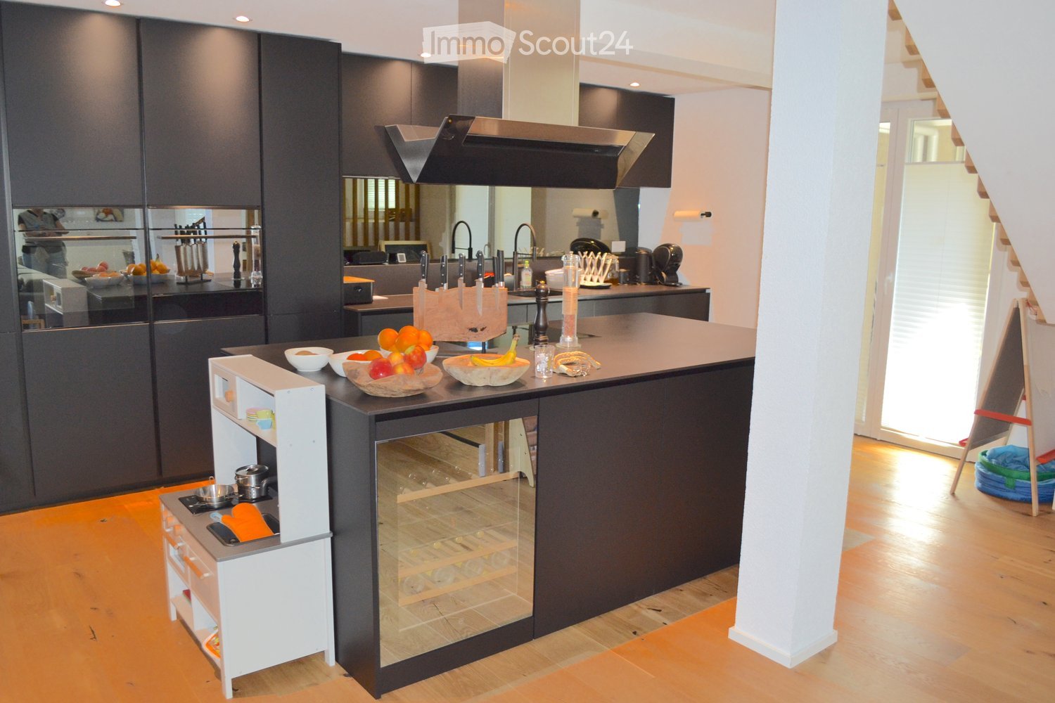 Modern kitchen with dark cabinets, a central island, and a built-in oven. The island features a wine rack, a microwave, and a small sink. The countertops display fruits and a knife block. The kitchen has a sleek, contemporary design with a wooden floor an