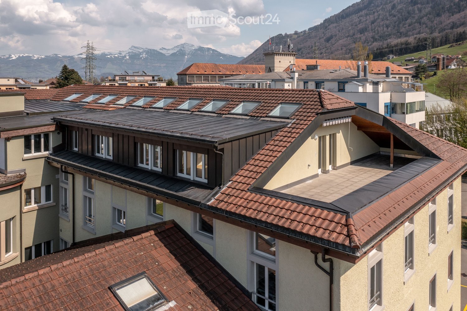 A multi-story residential building with red-tiled roofs, multiple windows, and a flat rooftop terrace. The surrounding area features other buildings and a scenic mountain backdrop.