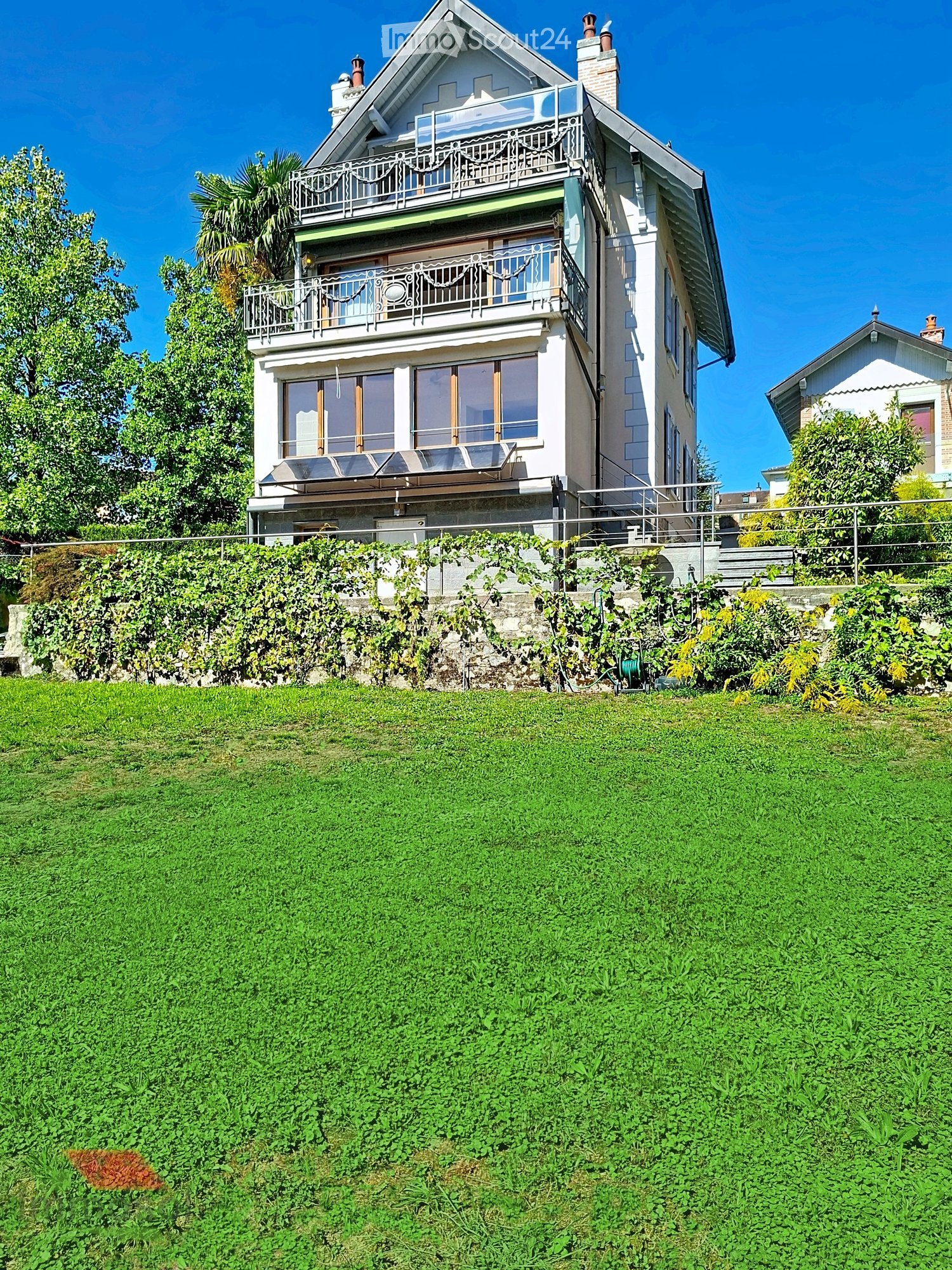 A two-story house with a raised ground floor, featuring balconies, large windows, and a well-maintained garden with green grass and climbing plants.