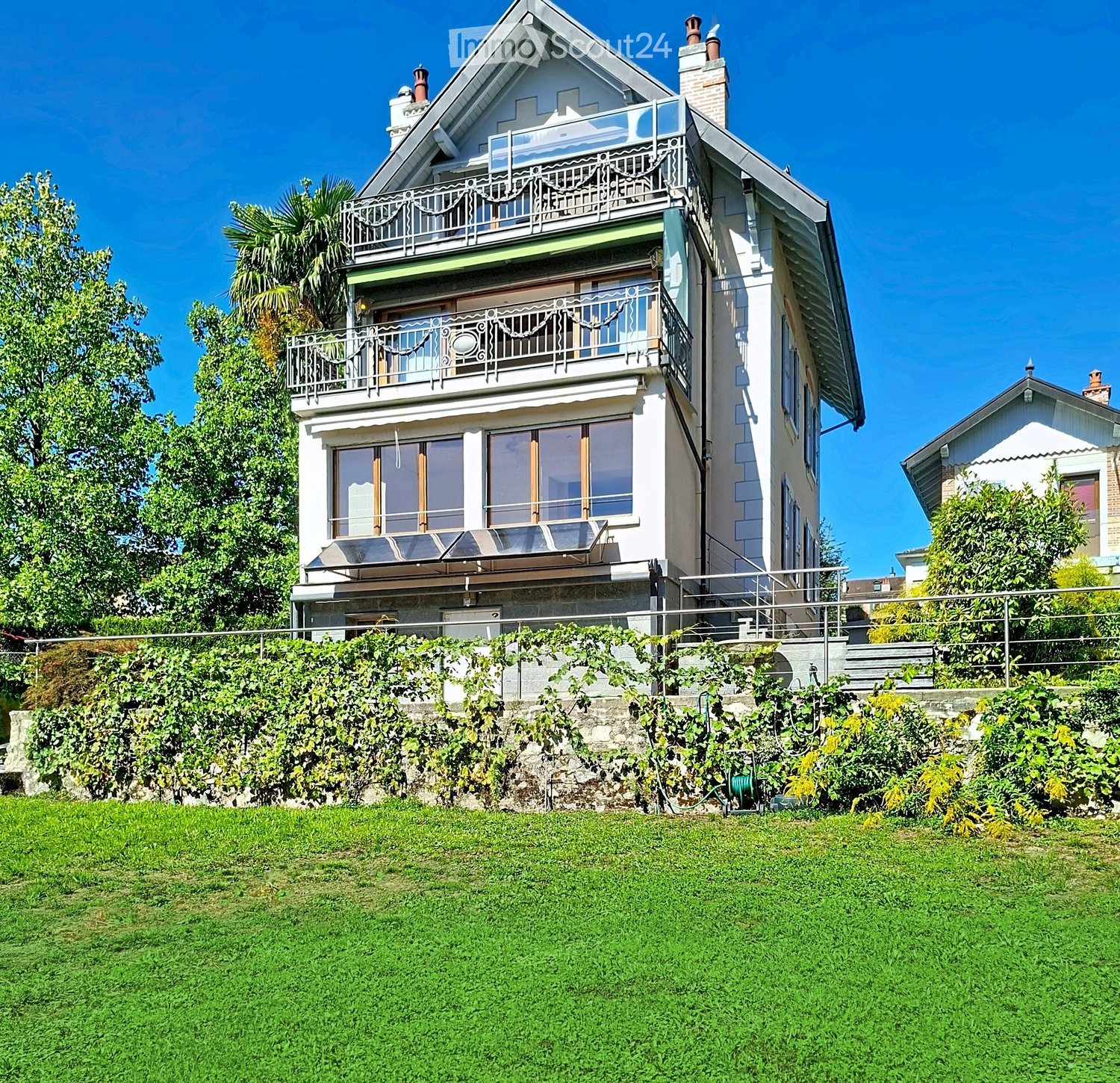 Two-story house with balconies, raised ground floor, and lush greenery surrounding the property.