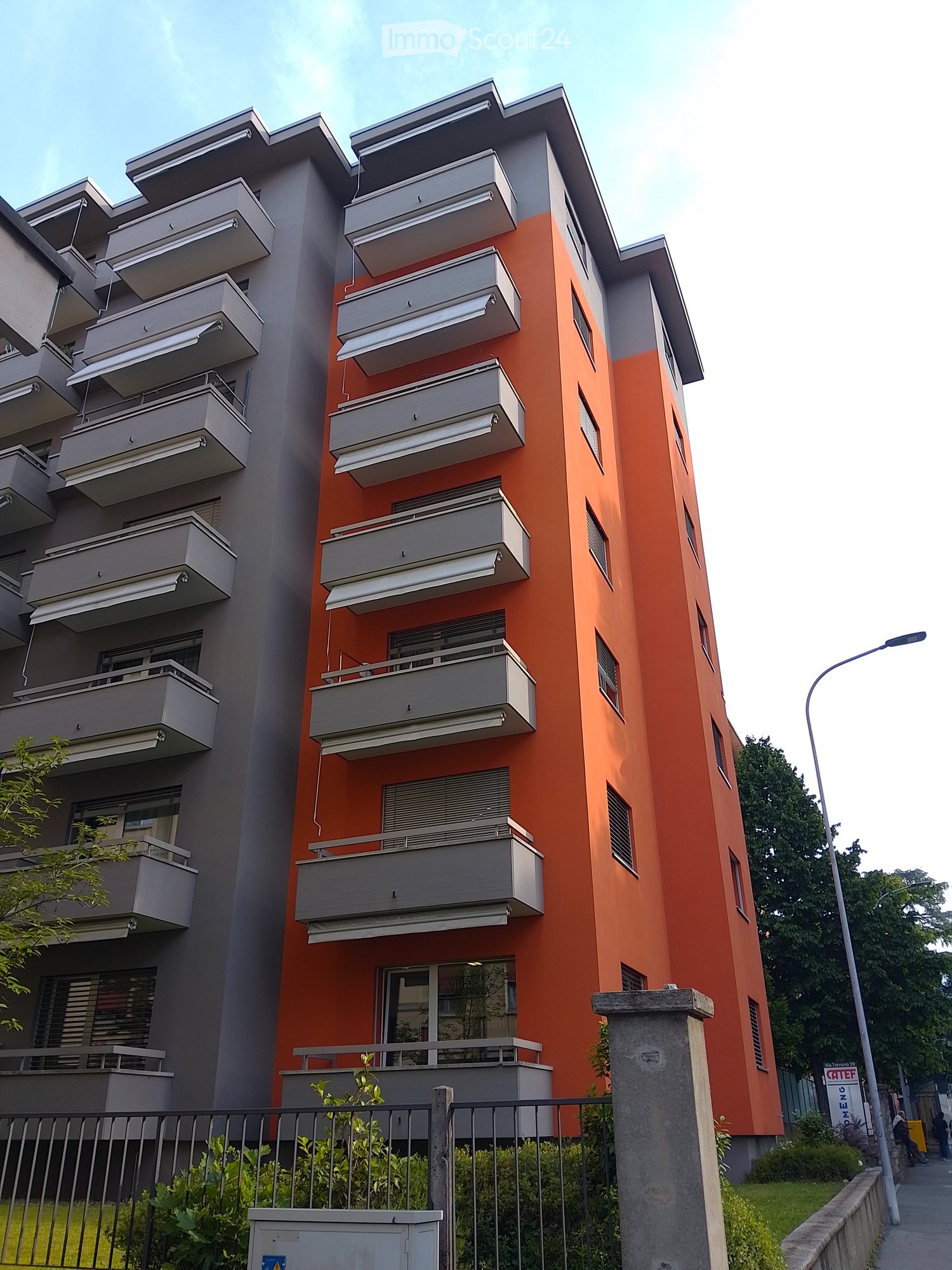 Modern multi-story apartment building with orange and grey facade, multiple balconies, and a gated entrance.