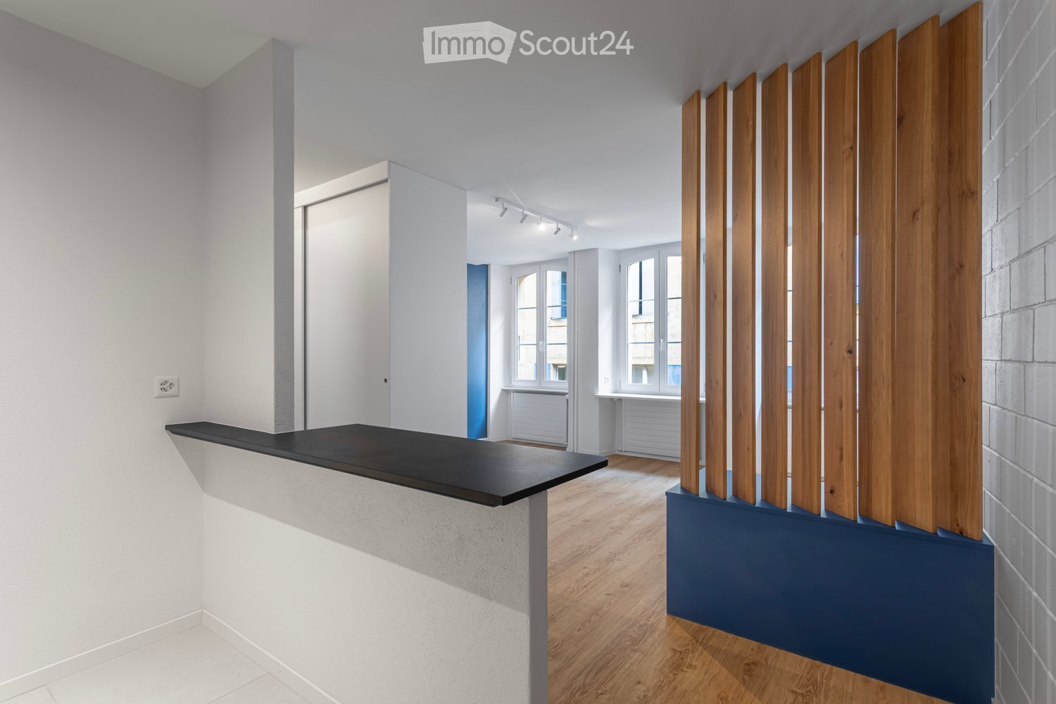 Empty room with white walls, wooden flooring, and a black countertop. The space features a modern design with large windows and a blue accent wall.