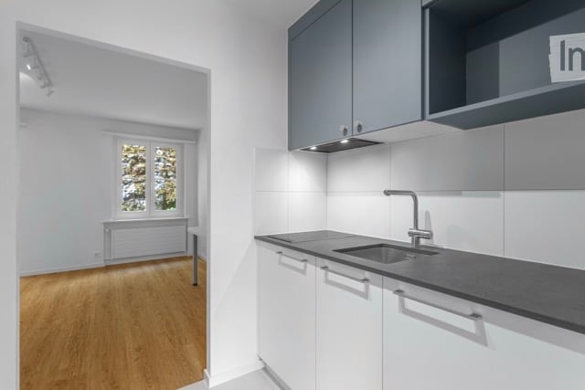 Empty room with wooden flooring, white walls, and a window with a view of trees. The adjacent kitchen area features white cabinets, a black countertop, and a sink.