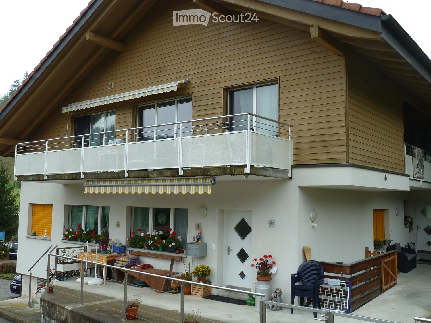 Two-story house with wooden facade, white lower level, balcony with metal railing, and outdoor seating area with chairs and table.