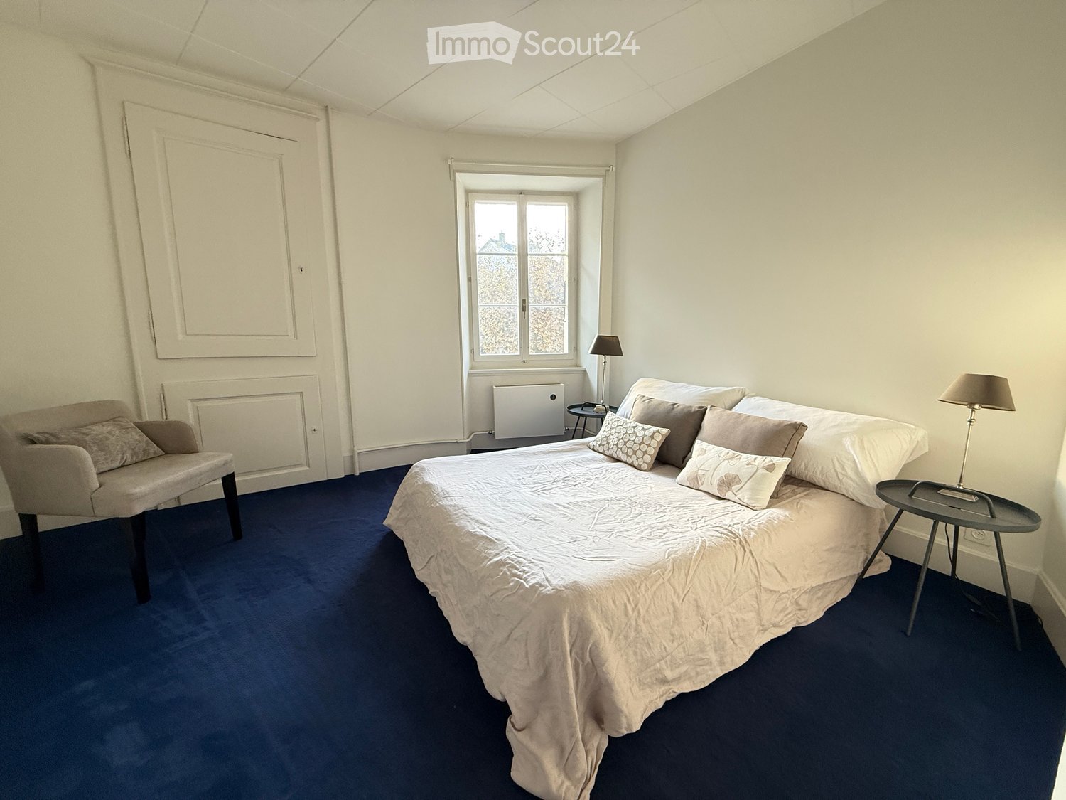 Cozy bedroom with a large bed, nightstands, a chair, and table lamps. The room features blue carpeting and white walls.