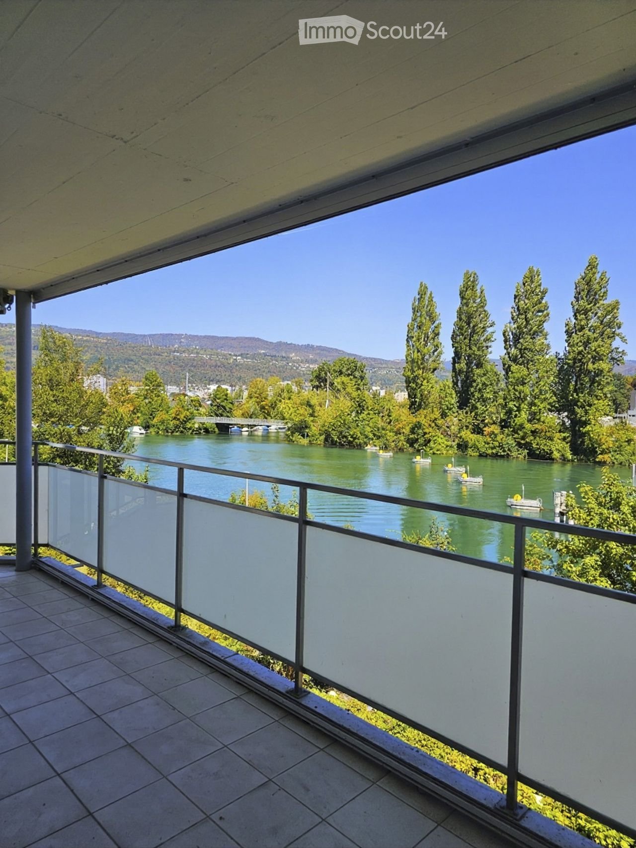 Balcony with a modern glass railing, overlooking a river with boats and surrounding greenery and hills.