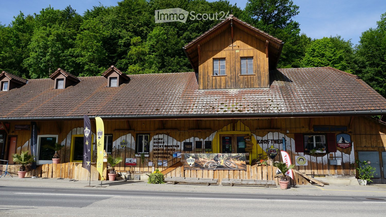 Two-story wooden building with a tiled roof, multiple windows, and a storefront with signage and banners. The building has a rustic design with wooden walls and a covered entrance area.