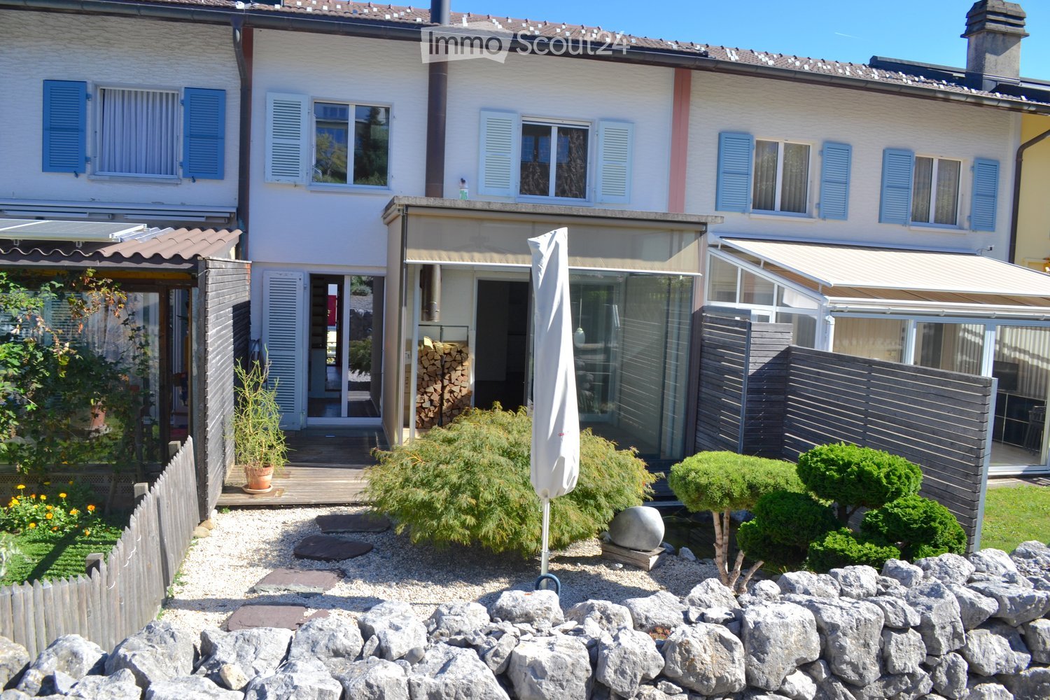 Two-story house with white walls, blue shutters, and multiple windows. The property includes a patio area with a wooden deck, outdoor furniture, and a well-maintained garden with gravel, plants, and a stone sculpture.