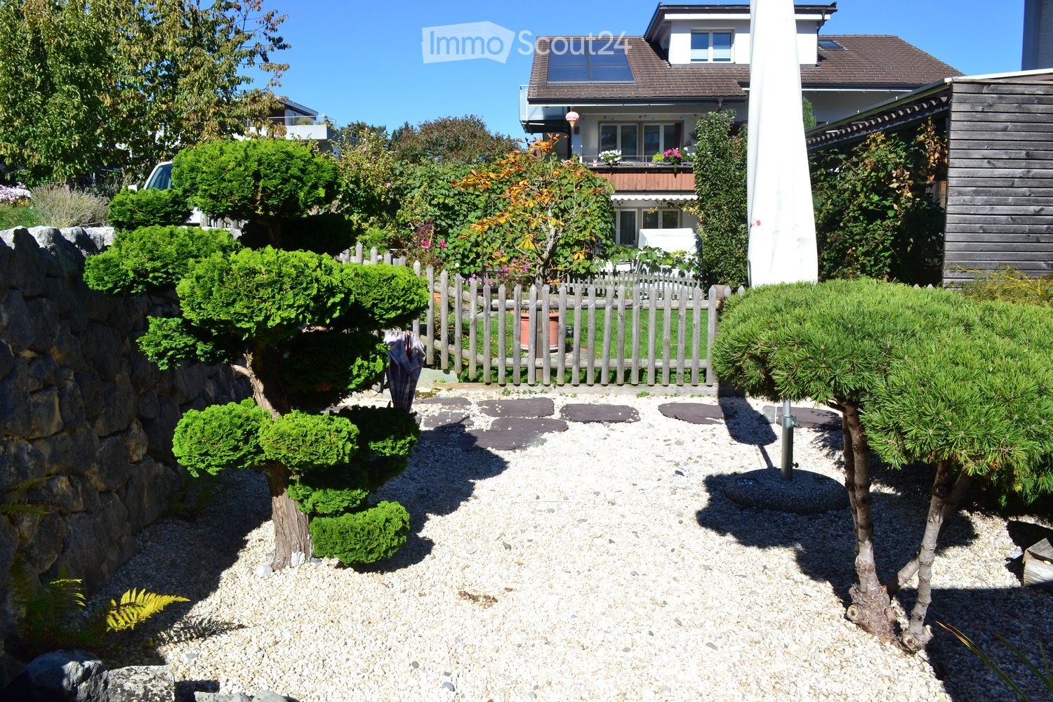 Well-maintained garden with gravel pathway, trimmed shrubs, small trees, stone border, wooden fence, and a house in the background.