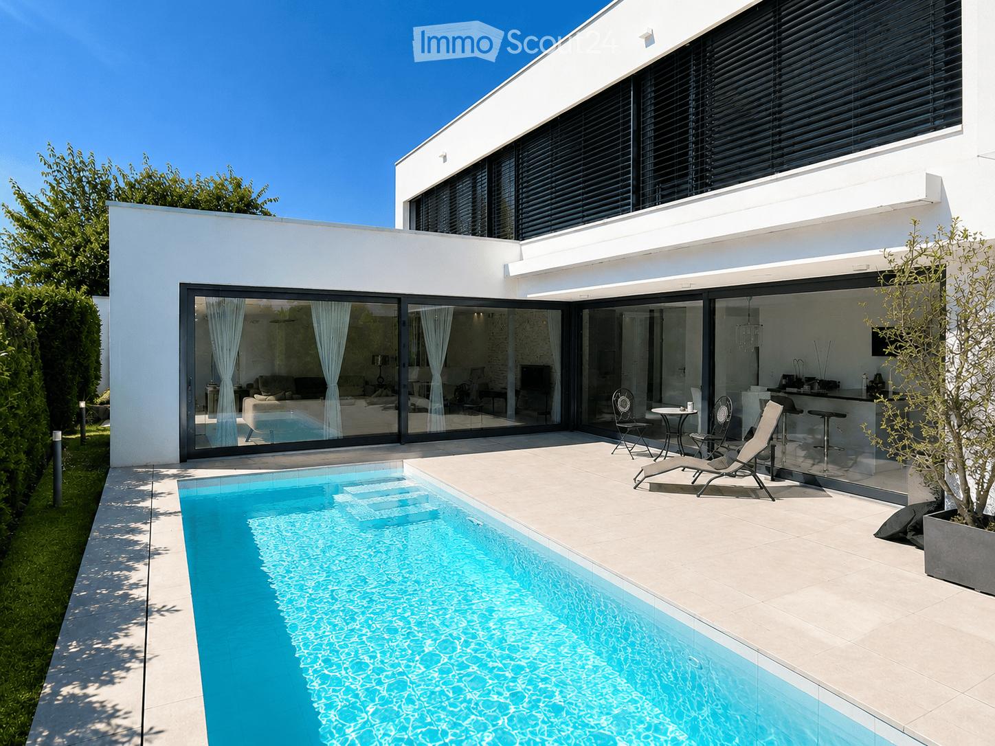 Modern two-story house with large glass sliding doors, a swimming pool, outdoor seating area with lounge chairs and a small table, and a well-maintained garden.