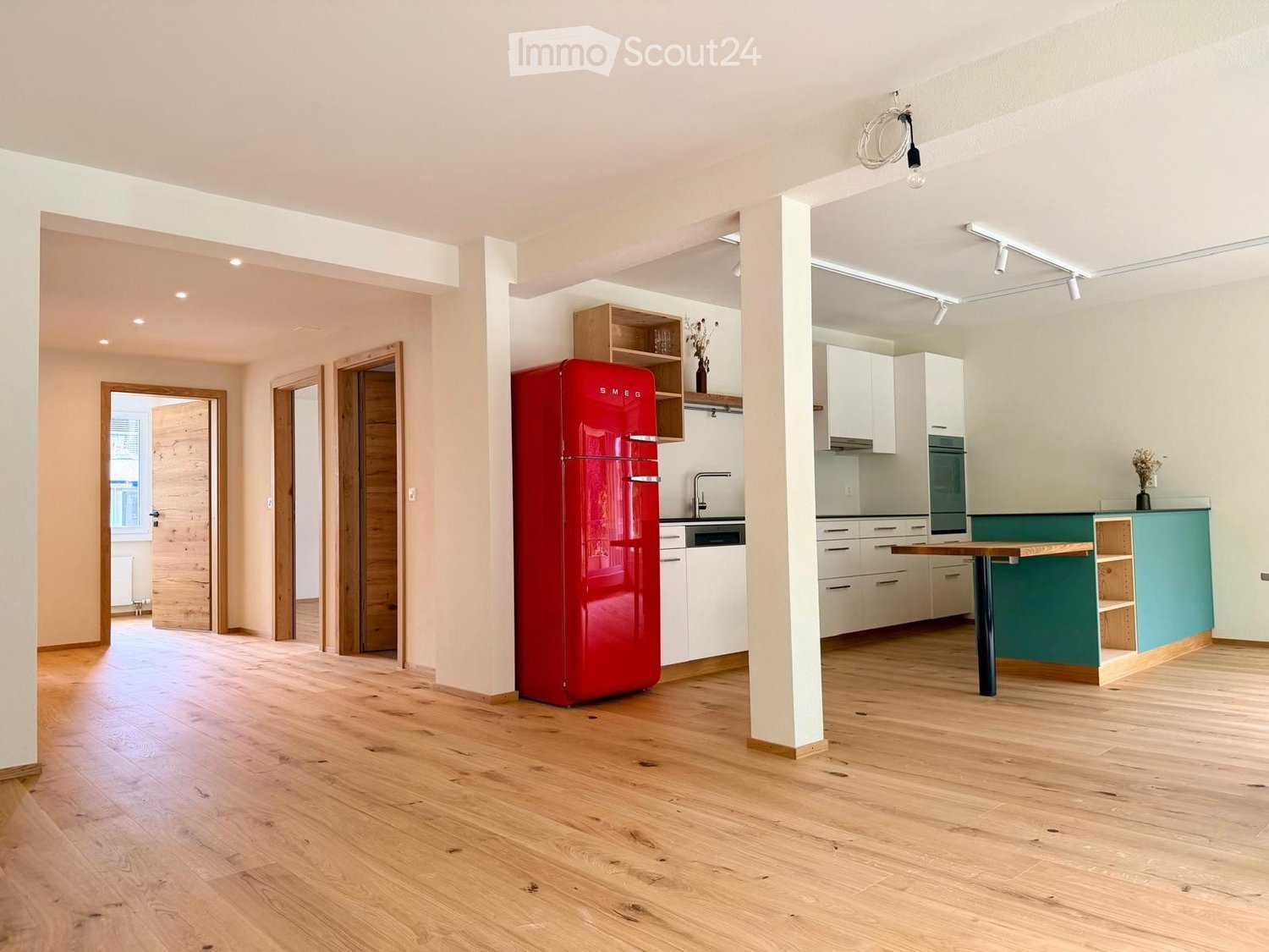 Open-plan kitchen and living area with light wooden flooring, white cabinets, a red refrigerator, a wooden table, and built-in shelving.