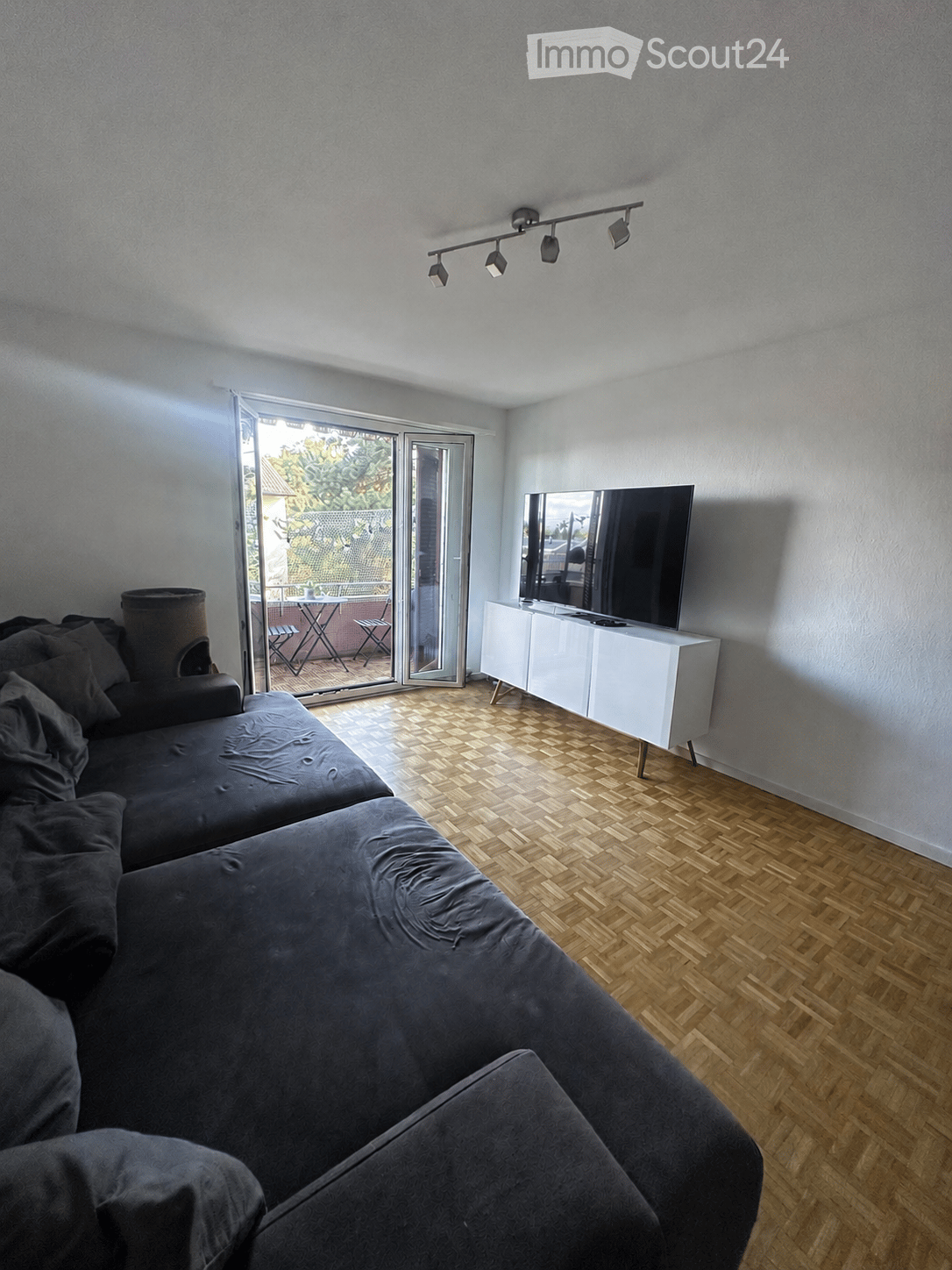 Modern living room with a grey sectional sofa, a flat-screen TV on a white cabinet, and a sliding glass door leading to a balcony with outdoor furniture.