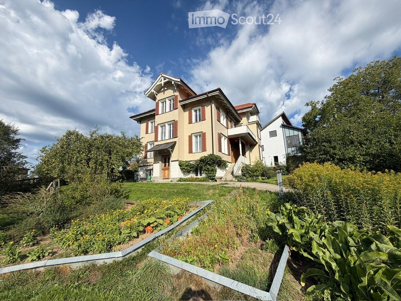 A two-story house with a beige facade, red shutters, and a well-maintained garden in front.