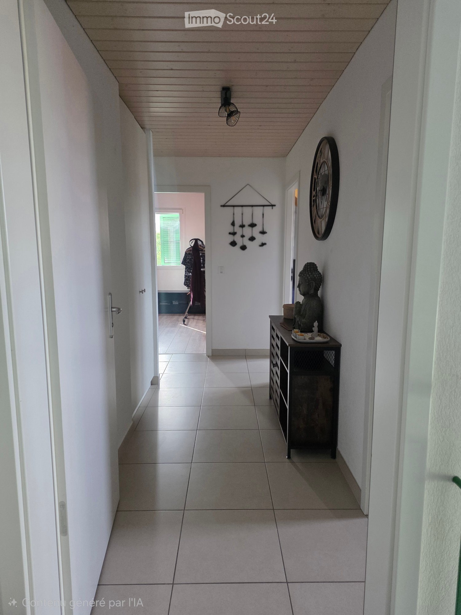 A clean, modern hallway with white tiled flooring, white walls, and a wooden ceiling. Features include a small table with a decorative Buddha statue, a wall-mounted clock, and a hanging wind chime. A doorway leads to another room with visible furniture.