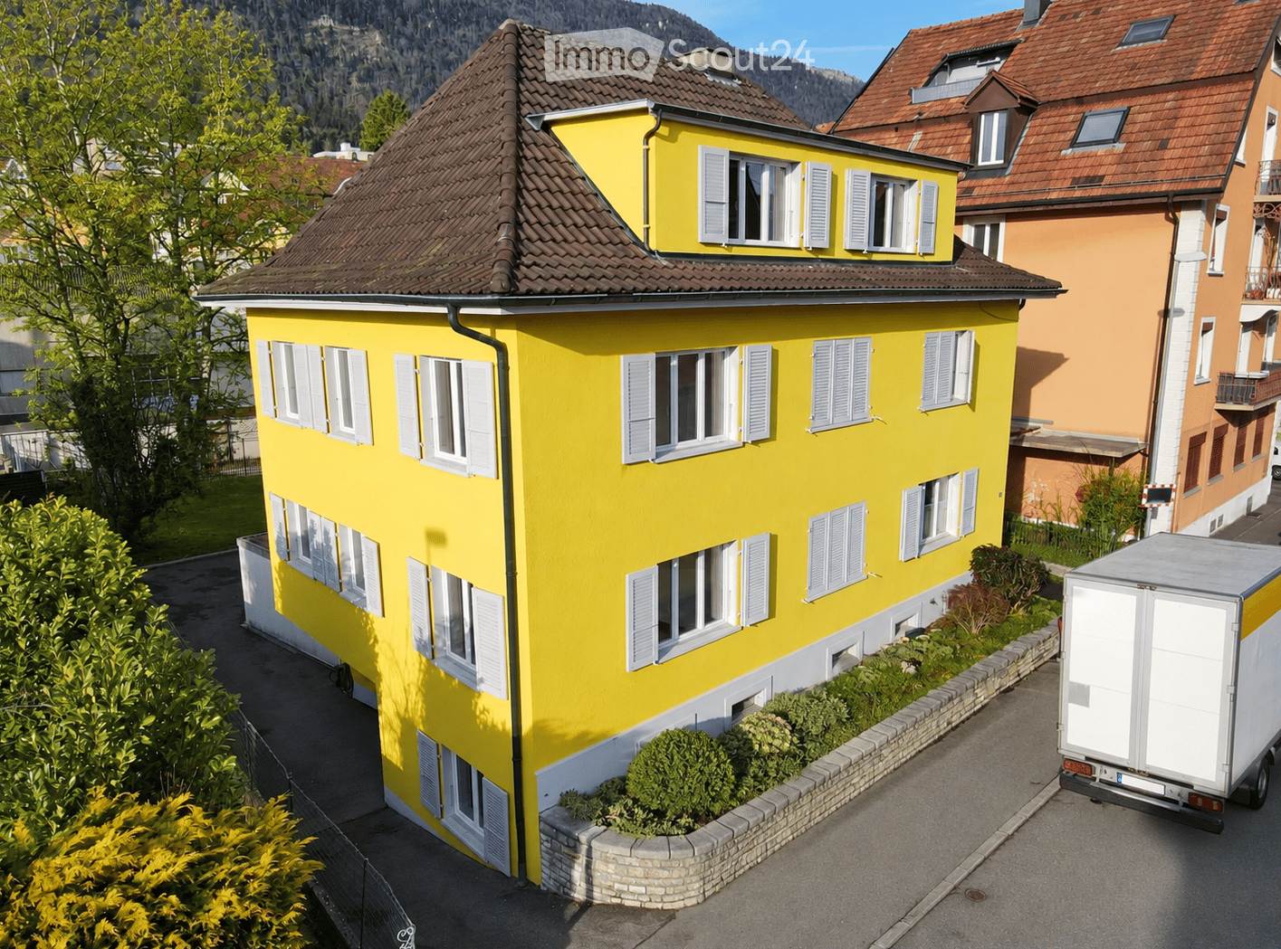 Bright yellow two-story house with white shutters and multiple windows, surrounded by greenery and a neighboring building.