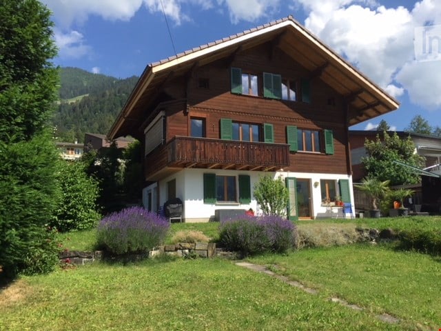 Two-story wooden house with green shutters, balcony, and well-maintained garden with lavender plants and a stone pathway.