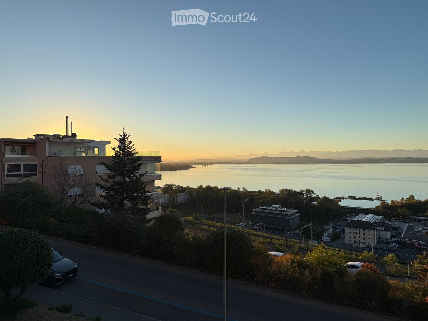 A modern multi-story building with balconies overlooking a serene lake and distant mountains during sunset.