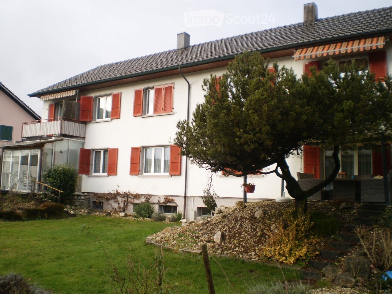 Two-story house with white walls, red shutters, and a tiled roof. Features include a balcony, garden, and a tree in the front yard.